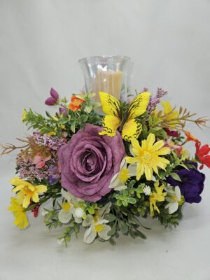 Handmade spring floral candle holder centerpiece with purple rose, colorful wildflowers, greenery, and yellow butterfly surrounding a glass hurricane candle holder.