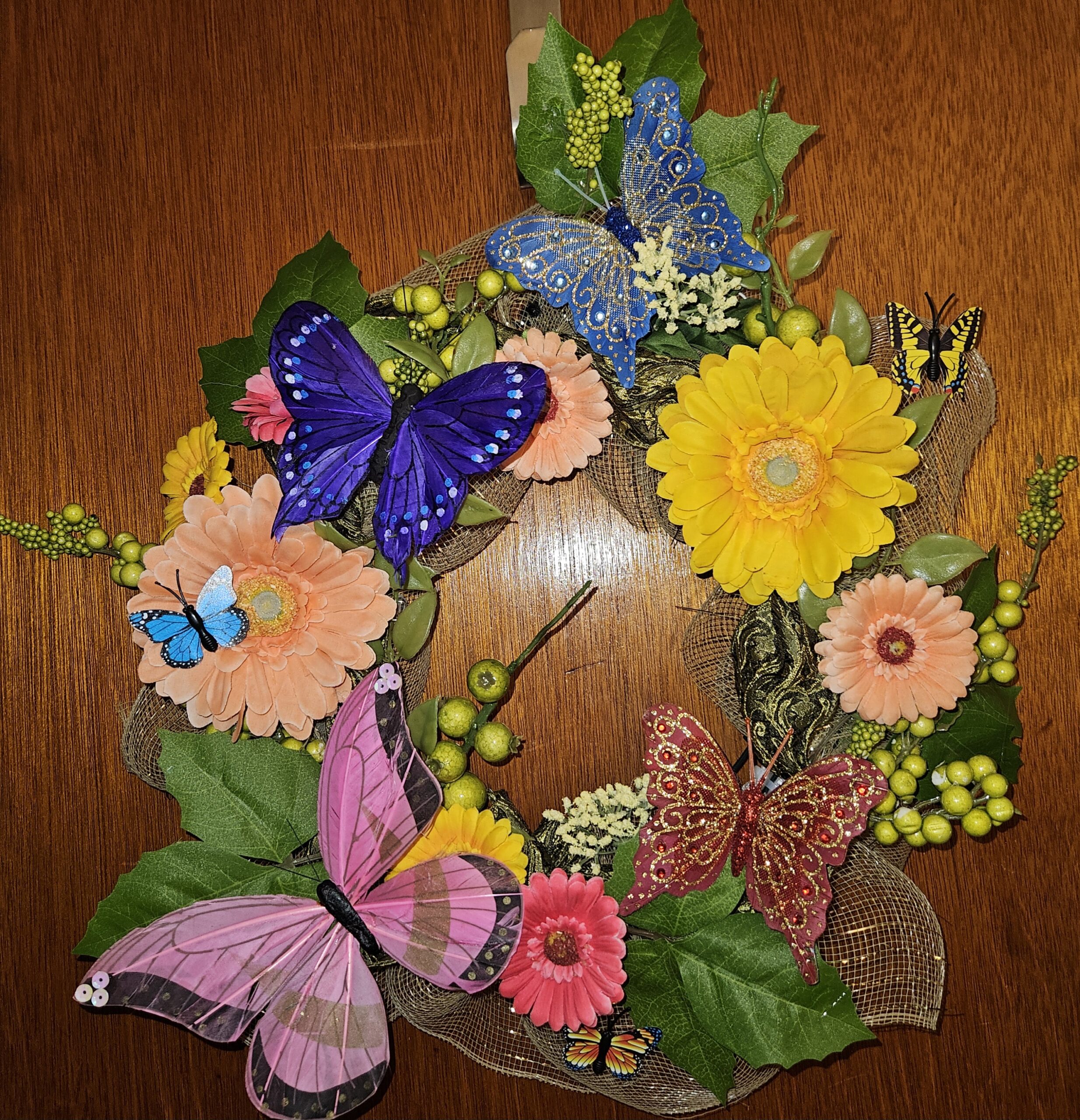 A vibrant handmade 21-inch wreath featuring yellow, peach, and pink gerbera daisies, lush green leaves, decorative berries, mesh ribbon, and large colorful butterflies in blue, purple, pink, and red, arranged in a full circular design for spring and summer decorating.