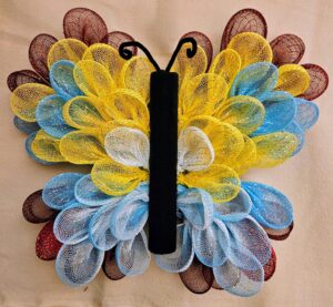 Handmade deco mesh butterfly wreath in vibrant yellow, blue, white, and maroon colors with black fleece body and curled antennae, 16x18 inches, displayed flat on a neutral background.