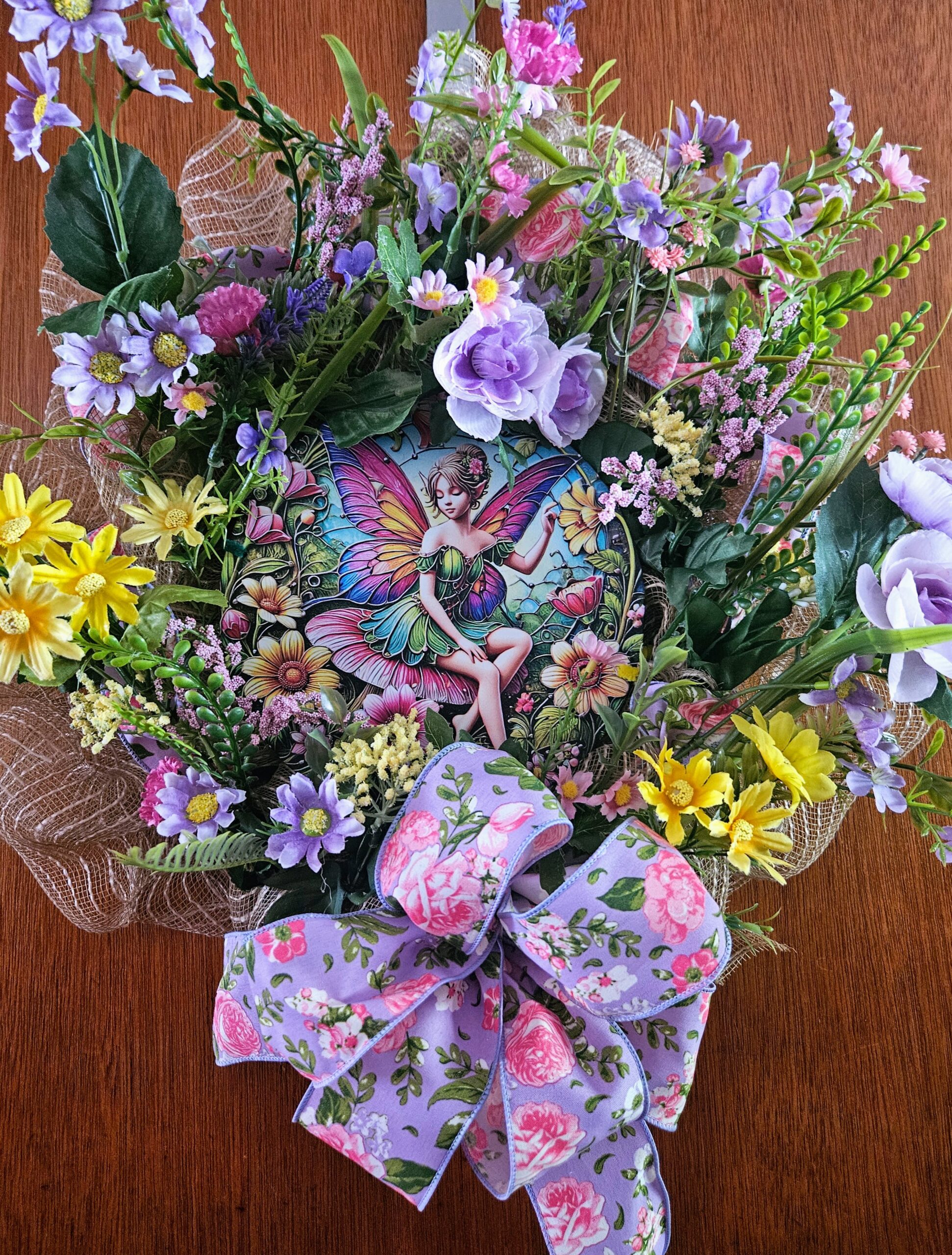 Large fairy garden wreath with purple flowers, daisies, greenery, and a central fairy illustration, finished with a lavender floral bow.