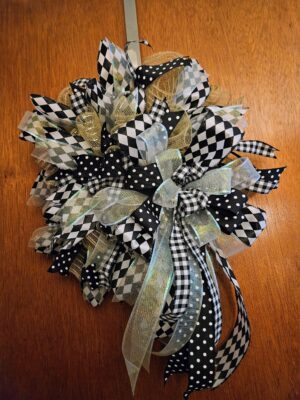 Close-up of a large handmade black and white mesh wreath featuring harlequin checks, polka dots, gingham, gold and iridescent mesh ribbons, and a dramatic center bow on a wooden door background.