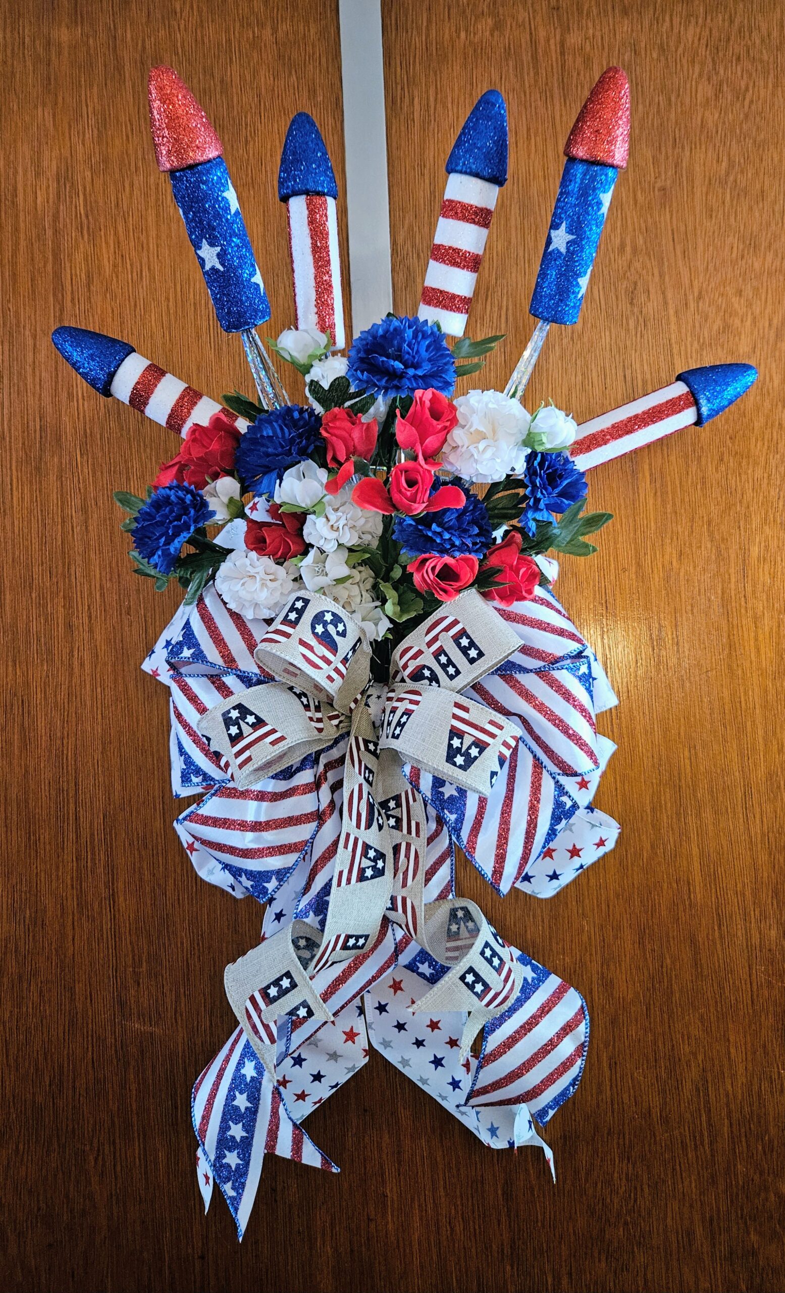 Handmade patriotic firework rocket swag featuring glitter rockets, red white and blue flowers, and festive Americana ribbons.