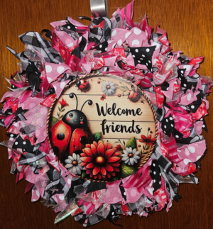 Handmade ladybug ribbon wreath featuring pink patterned ribbons and a Welcome Friends ladybug sign with flowers.